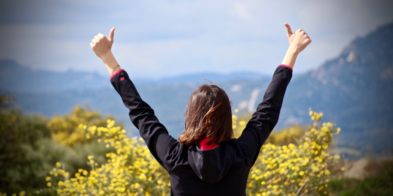 A person in a black shirt holds two thumbs up in the air with their back turned facing a mountain scene. 