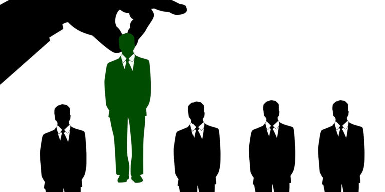 A line of black silhouette of people in suits and ties with one colored green and being picked up by a large hand.