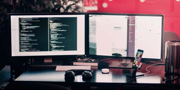 Two computer monitors side by side showing code on the left and software applications on the right with headphones and an iPhone in front.