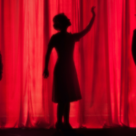 The silhouette of a  woman sings while two men on either side of her act out their roles with a red theater backdrop.