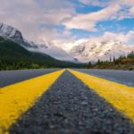 Two yellow lines come together to point down the road towards a mountain and cloudy skies