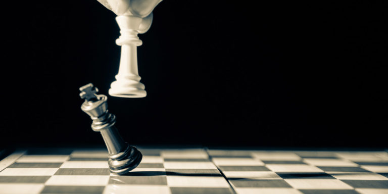 A hand guides a white queen chess piece and knocks over a black king chess piece on a black and white chessboard.