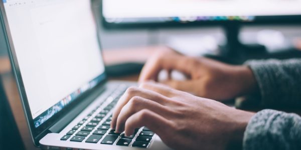 A person types on a laptop with a computer monitor in the background.