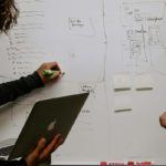 Two people stand in front of a busy whiteboard one is watching while the other writes and holds a laptop.