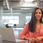 A woman stands at desk and looks at the camera with a smile.