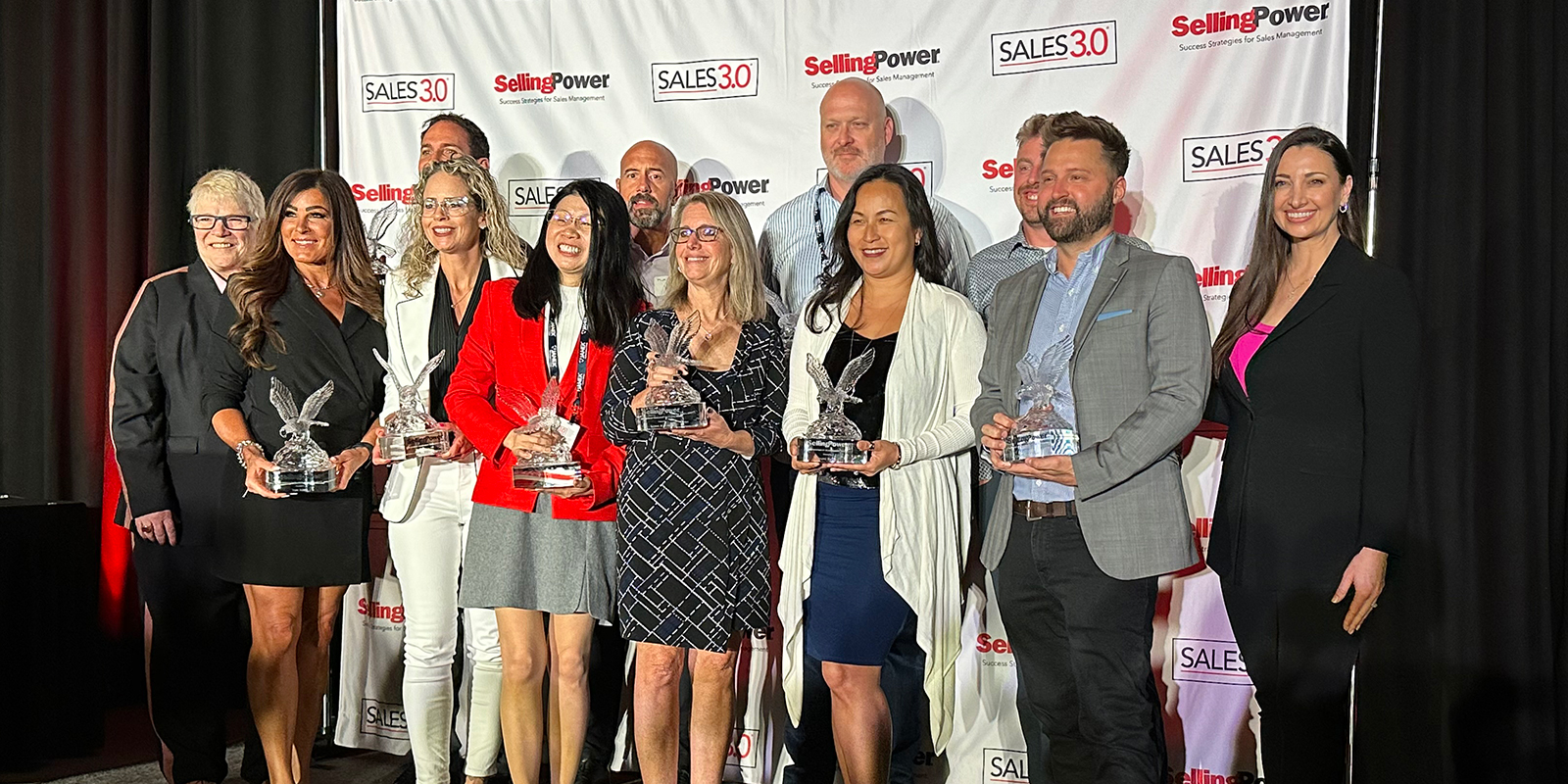 Twelve people stand in a group smiling and holding awards.