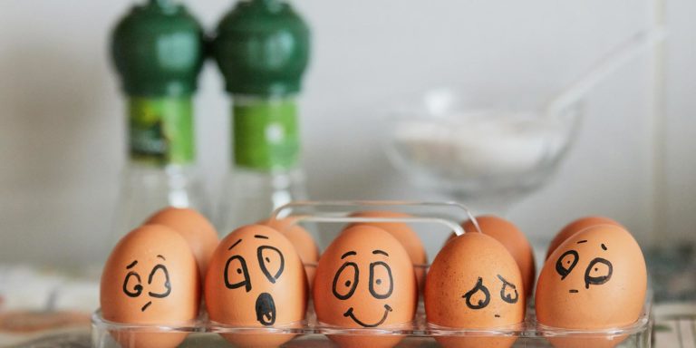 Five brown eggs sit in a carton each with different facial expressions drawn on them.