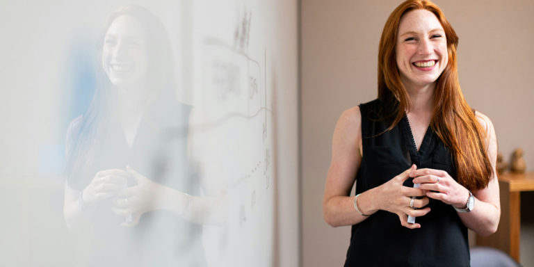 A woman stands in front of a whiteboard looking off camera.