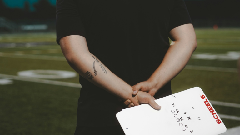 A football coach holds a whiteboard behind his back on the football field.